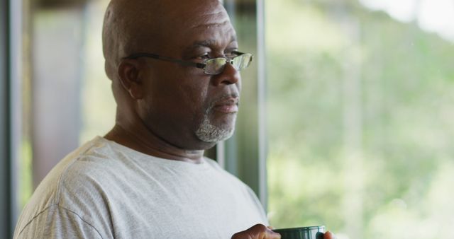 Elderly man stands alone near window, holding mug with thoughtful expression. Natural light illuminates his face, creating a serene atmosphere. Ideal for themes related to aging, contemplation, morning routines, and solitary moments.