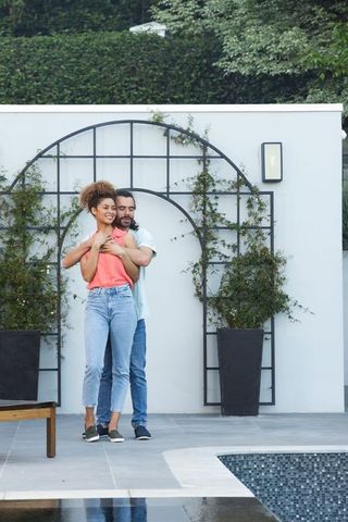 Mixed-race couple embracing poolside on modern patio with trellis and lush garden backdrop