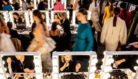 Backstage makeup artists preparing models at lighted vanity mirrors for high-fashion runway