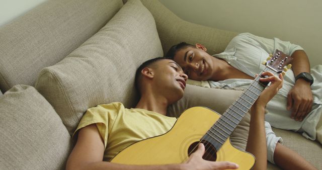 Happy biracial gay male couple lying on sofa one playing guitar and singing. staying at home in isolation during quarantine lockdown.