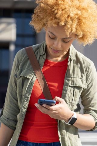 African american woman checking smartphone outdoors in sunny city with curly blonde hair