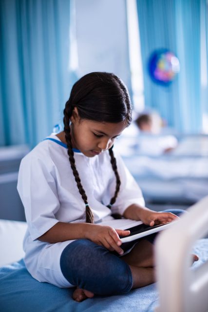 Young girl sitting on hospital bed using digital tablet. Ideal for illustrating pediatric healthcare, modern medical technology, patient care, and hospital environments. Useful for healthcare websites, medical brochures, and educational materials about child patients and hospital stays.