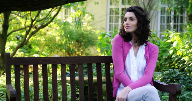 Young woman with dark curly hair in pink cardigan and white pants sitting on wooden bench in lush garden, looking contemplative. Ideal for themes of tranquility, relaxation, nature, outdoor enjoyment, lifestyle, and peaceful settings.