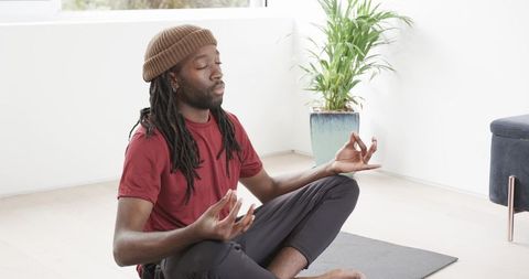 African American man meditating on yoga mat wearing beanie in bright minimalist room