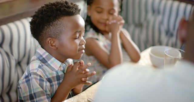 Children sitting together at restaurant table praying before meal. Illustrates gratitude and faith, perfect for family-focused content, religious publications, and lifestyle promotions.