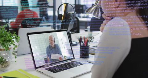 Senior consultant mentoring remotely over laptop with code overlay in modern open office