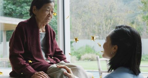 Caregiver comforting senior by window holding knee wearing burgundy cardigan with gold stars