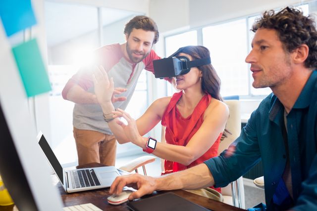 Colleagues in a modern office are collaborating using virtual reality technology. One person is wearing a VR headset while others are engaged, indicating teamwork and innovation. This image is ideal for illustrating concepts related to technology, digital transformation, and collaborative work environments.