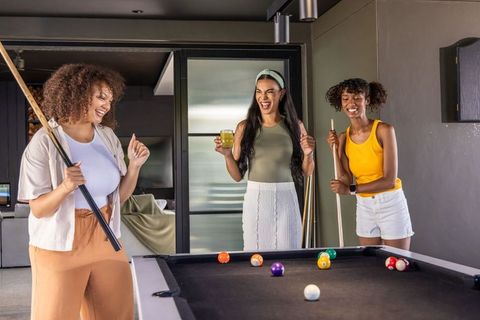 Diverse women playing pool and laughing while enjoying drinks in stylish home game room