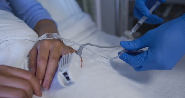 Healthcare professional in blue gloves administering an IV to patient’s hand in a hospital bed. Useful for illustrating healthcare services, hospital environments, patient care, or medical treatments.