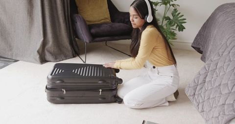 Young woman packing suitcase on bedroom floor wearing headphones preparing for travel