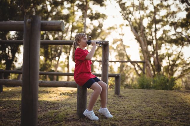Young girl in red shirt and black shorts drinking water after completing an obstacle course in a boot camp. She is sitting on a wooden structure in an outdoor setting with trees in the background. Ideal for use in content related to children's fitness, outdoor activities, hydration, and healthy lifestyles.