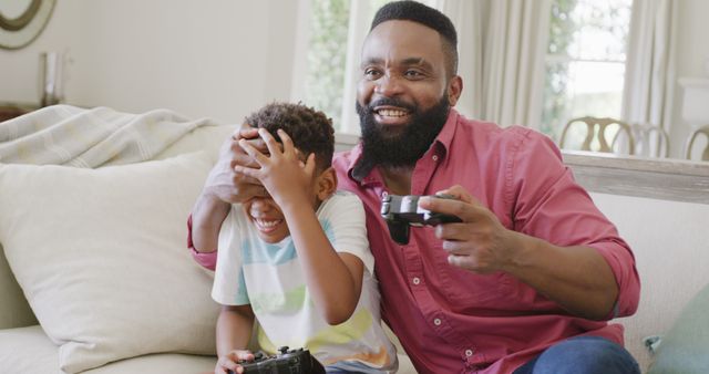 Father and son playing video games together in living room, father showing excitement while son covers laughter. Perfect for themes around family bonding, father-son relationship, technology at home, domestic life, happiness, and leisure activities.