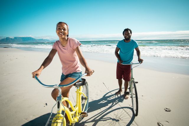 African American couple enjoying a sunny day at the beach, wheeling their bicycles along the shoreline. Ideal for promoting summer holidays, romantic getaways, outdoor activities, and healthy lifestyles. Perfect for travel brochures, lifestyle blogs, and advertisements focused on leisure and recreation.