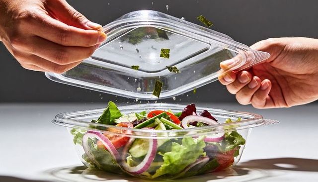 Hands lifting clear lid over fresh mixed greens salad in plastic clamshell, herbs falling