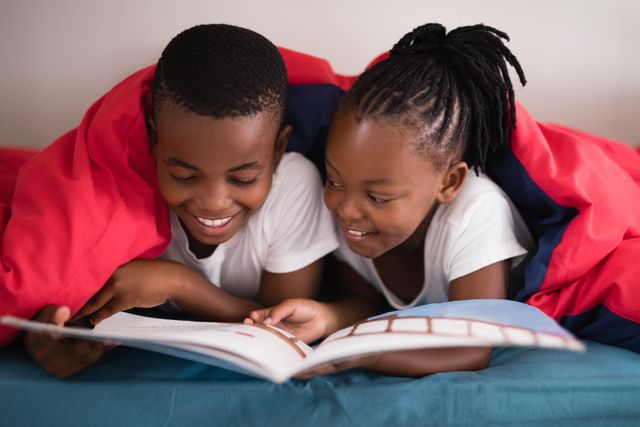 Smiling siblings reading book together while lying on bed at home