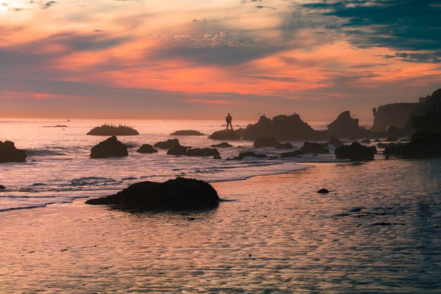 This photo shows a solitary figure standing on rocky shore during a beautiful sunset. Ideal for concepts of peace, solitude, natural serenity, and scenic landscapes. Perfect for travel blogs, nature publications, and websites featuring calm and tranquil scenes.