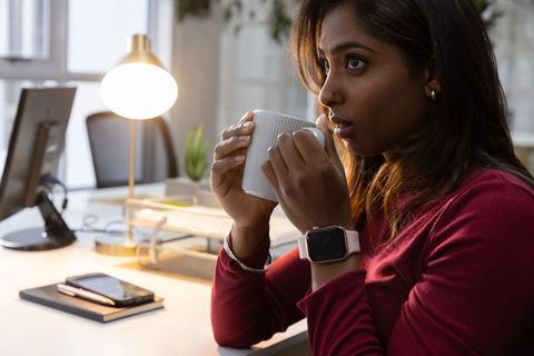 Contemplative indian woman sipping coffee at minimalist office desk with smartwatch