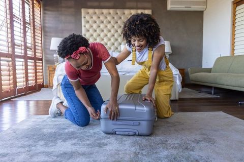Laughing while packing: black women kneeling on rug closing suitcase for weekend trip