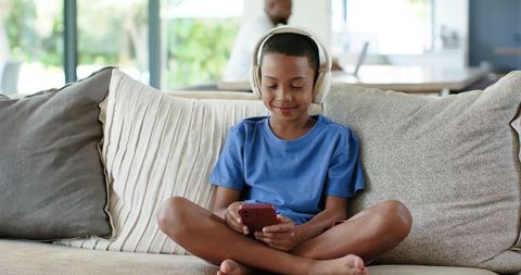African american boy listening with headphones and using smartphone on living room sofa