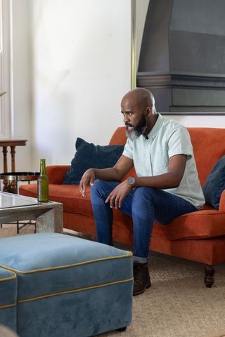 Mature african american man sitting on orange sofa looking intently with green bottle nearby