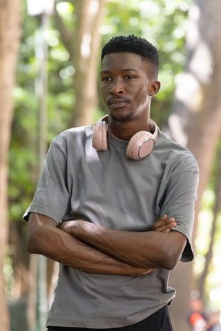 Contemplative african man standing with arms crossed wearing pink headphones in green park