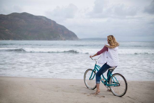 Mature woman riding bicycle along sandy beach with ocean waves in background. Ideal for promoting outdoor activities, healthy lifestyle, travel destinations, and leisure time. Perfect for use in advertisements, travel brochures, wellness blogs, and lifestyle magazines.