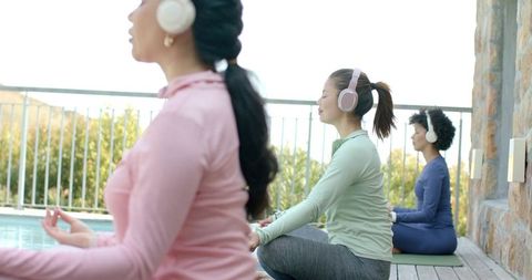 Diverse women meditating poolside with wireless headphones on sunlit terrace