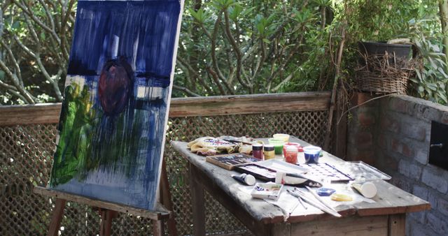 This image showcases an outdoor art studio with an abstract painting on an easel. The workspace has a table filled with colors, brushes, and other art supplies, surrounded by garden greenery. Perfect for articles about outdoor creativity, artist workspace setups, painting classes in nature, and promoting tranquility in creative pursuits.