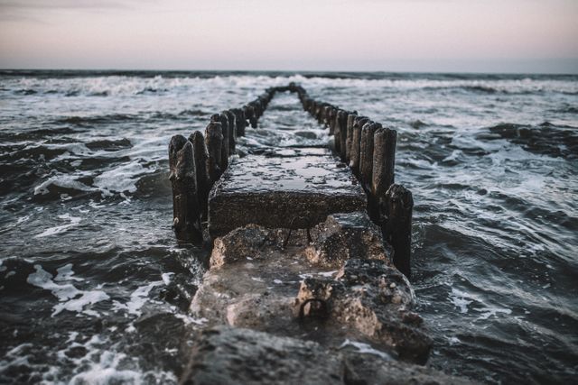 An abandoned jetty dramatically extends into the rough sea with powerful waves crashing in. The scene is captured at sunset, with a moody sky and turbulent water creating a dramatic atmosphere. This image is perfect for use in articles related to coastal life, environmental issues, photography focused on natural landscapes, and relaxation or meditation content emphasizing serene and solitary sceneries.