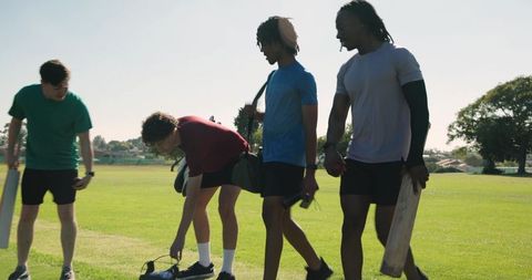 Heading to cricket match carrying bats, diverse male friends walking on sunlit sports field