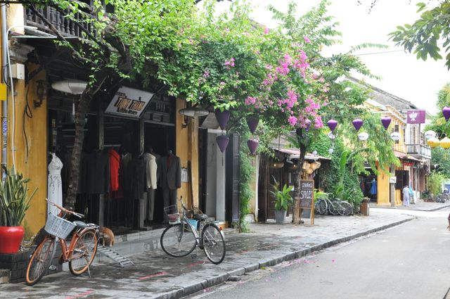 Colorful scene of Hoi An showcases typical Vietnamese architecture with charming clothing shops, flowers, and classic bicycles. Suitable for travel magazines, promotional materials, and blogs about travel destinations in Vietnam. Captures serene, picturesque street ideal for promoting tourism.