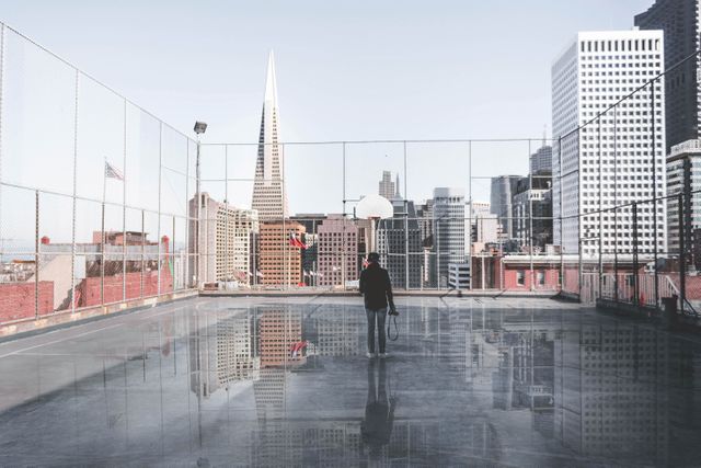 Lone person stands on an urban rooftop basketball court, holding basketball with city skyline in background. Suitable for illustrating urban sports, solitude in the city, urban recreation, city lifestyle, athletic activities, modern city life, and reflections.