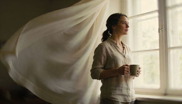 Woman standing at window holding mug, gazing contemplatively, billowing curtain, soft light