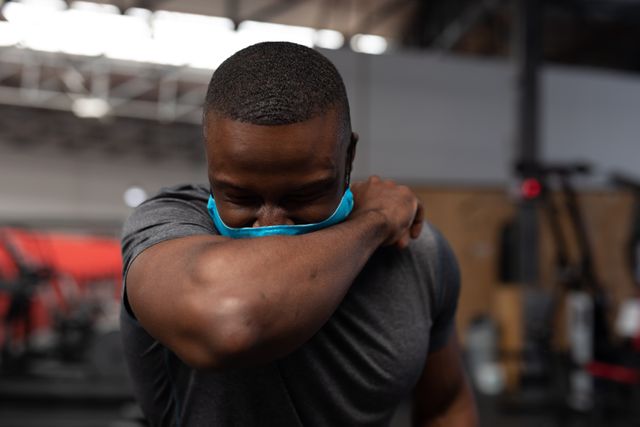 African American man with toned muscles covering his mouth and nose with his arm while sneezing in a gym. He is wearing a blue face mask, emphasizing health and hygiene during workouts. Useful for topics related to fitness, health safety, pandemic precautions, and gym environments.