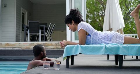 Poolside couple chatting and relaxing on deck lounger with refreshing drinks