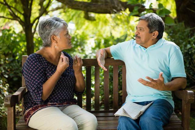 Senior couple sitting on a wooden bench in a park, engaging in a serious conversation. The setting is lush with greenery, indicating a peaceful outdoor environment. This image can be used for topics related to elderly relationships, communication, retirement life, and outdoor activities for seniors.