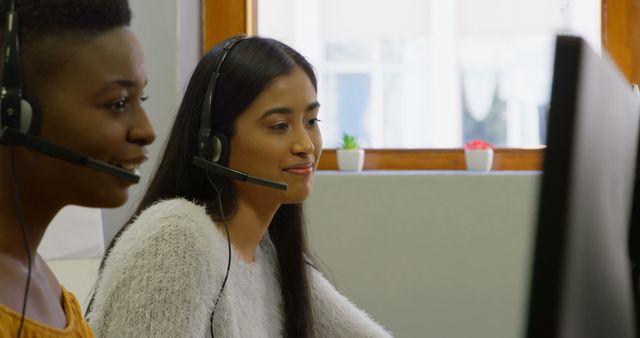 Two female customer service representatives are working at their desks with headsets, showing a diverse work environment. This can be used for content related to customer support, teamwork, office environment, and professional service settings.