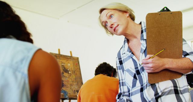 Art instructor observing students working on their paintings in classroom. The teacher, holding a clipboard and pencil, provides guidance and critiques to enhance the students' creativity and skills. Useful for educational materials, art school advertisements, and creativity tutorials.