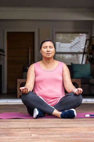 Mature asian woman meditating on home deck doorway wearing sportswear and earbuds