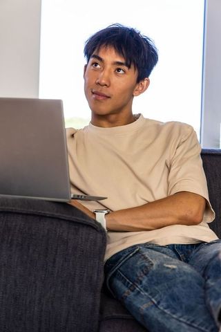 Young asian man working remotely on laptop from couch, wearing smartwatch, casual home