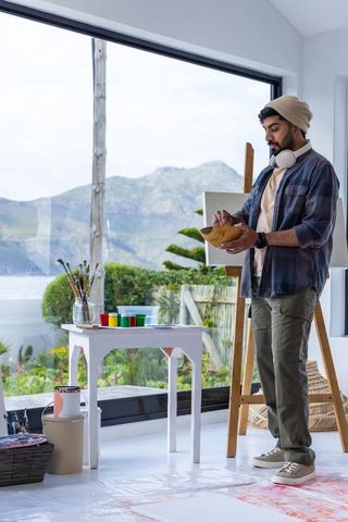 Indian artist mixing paint in sunlit coastal studio with ocean view and mountain backdrop