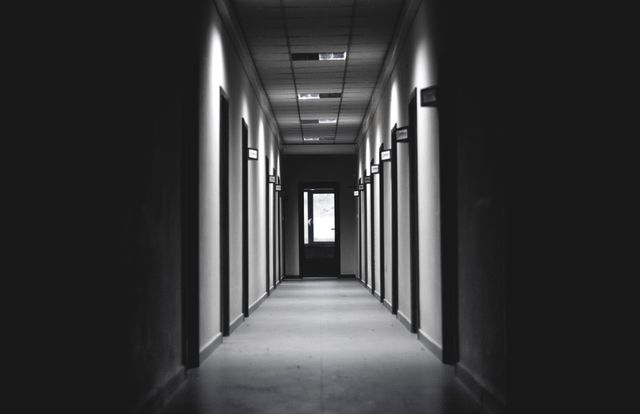 Dimly lit hallway in office building with closed doors on both sides creates eerie, mysterious atmosphere. Ideal for illustrating themes related to mystery, suspense, loneliness, or abandoned places. Suitable for use in horror film scenes, novel covers, or psychological thriller promotions.