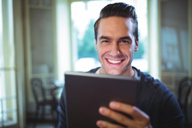 This image features a man smiling while using a digital tablet in a cafe. Ideal for promoting technology usage, casual lifestyle, or modern cafes. Useful for websites, blogs, and advertisements focused on tech, leisure, or social settings.
