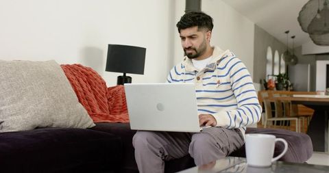 Indian man working on laptop on sofa in modern minimalist open-plan living room telework