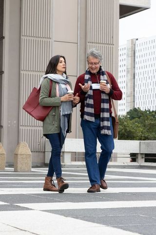 Mature couple walking and holding coffee cups on urban plaza with striped scarf and tote
