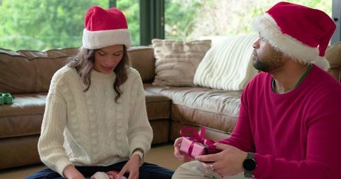 Couple exchanging christmas gifts on living room floor wearing santa hats holiday moment