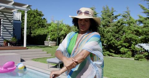 African american woman applying sunscreen by pool wearing straw hat and striped cover-up