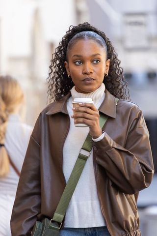 African american woman holding takeaway coffee, wearing brown leather jacket, urban street
