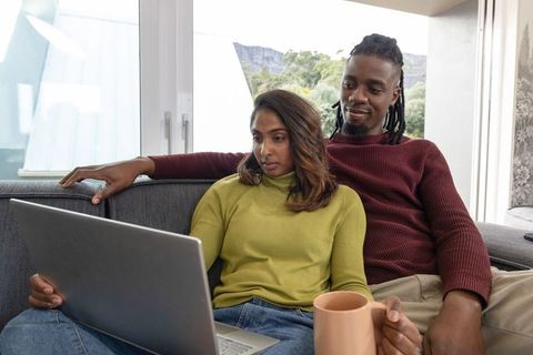 Mixed couple relaxing on sofa, browsing laptop and sharing coffee in bright living room